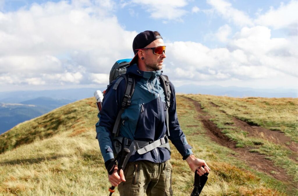 Man wearing polarized sunglasses and hiking gear on a mountain trail, with bright skies and distant hills in the background.